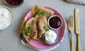 Glutenfria pannkakor med sylt och grädde på en rosa tallrik. En gaffel och kniv ligger bredvid.