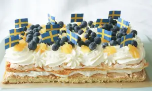 En stor brittatårta i långpanna, dekorerad med grädde, blåbär, apelsinbitar och små svenska flaggor. Perfekt för nationaldagsfirandet!