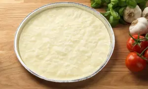 En rund, ljus deg i en folieform, redo för ett glutenfri pizzadeg recept. Tomater och vitlök i bakgrunden.
