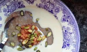 En skål med skrädmjölsgröt, toppad med äppelmos och pumpafrön, serverad i en vacker lila-vit tallrik.
