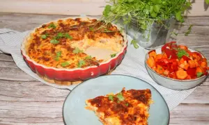 En gyllene paj med keso och en bit serverad på en tallrik. Bredvid står en skål med grönsaker och en kruka med örter.
