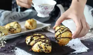 En hand plockar upp en chokladscone, dekorerad med chokladglasyr. Fler chokladscones ligger på ett fat.