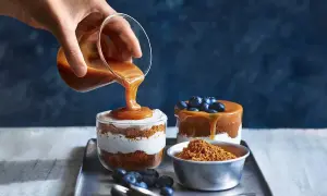 En hand häller karamellsås över en läcker cheesecake i glas. Blåbär och smulor fulländar desserten.