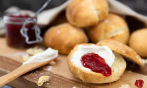 En sked med grädde och sylt på en halverad scones, redo att avnjutas. En burk sylt och fler av dessa bästa scones i bakgrunden.