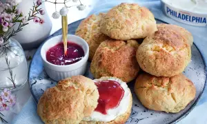 Färska scones bikarbonat på ett fat med sylt och grädde. En vacker bukett blommor i bakgrunden.