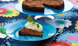 En bit kladdkaka med vispad grädde och mynta på en blå tallrik. Hela kladdkakan syns på en tårtfat i bakgrunden. Ett perfekt kladdkaka recept för fikat.