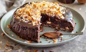 Kladdkaka med kokostosca, täckt med chokladglasyr och rostad kokos. En bit är uppskuren och visar den saftiga insidan.