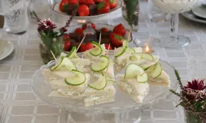 Små smörgåsar med gurka och gräddost, redo för afternoon tea. Jordgubbar och choklad i bakgrunden.