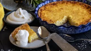 En gyllene ananaspaj med en bit serverad på en tallrik med vispad grädde. Bredvid står en skål med mer grädde och en hel ananas. Perfekt för ett ananaspaj recept.
