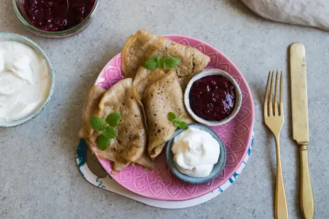 Glutenfria pannkakor med sylt och grädde på en rosa tallrik. En gaffel och kniv ligger bredvid.