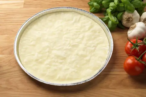 En rund, ljus deg i en folieform, redo för ett glutenfri pizzadeg recept. Tomater och vitlök i bakgrunden.
