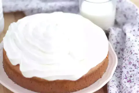 En saftig tres leches-kaka med fluffigt vitt täcke, bredvid ett glas mjölk och en blommig duk.