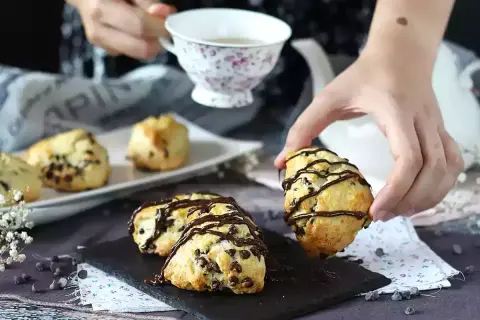 En hand plockar upp en chokladscone, dekorerad med chokladglasyr. Fler chokladscones ligger på ett fat.