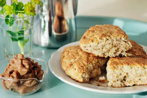 Nybakade kardemumma scones på en vit tallrik, med en skål fylld med kardemummafyllning och en vas med gröna blommor i bakgrunden.