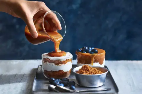 En hand häller karamellsås över en läcker cheesecake i glas. Blåbär och smulor fulländar desserten.