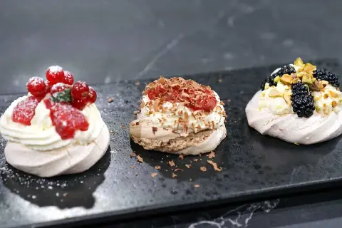 Tre små pavlovakakor med olika toppingar: jordgubbar och vinbär, choklad och hallon, samt björnbär och pistagenötter.