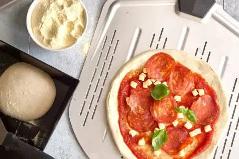 Pizzadeg recept: färdig pizzadeg med salami, ost och basilika på en pizzaspade. Bredvid ligger mer deg och en skål med mjöl.