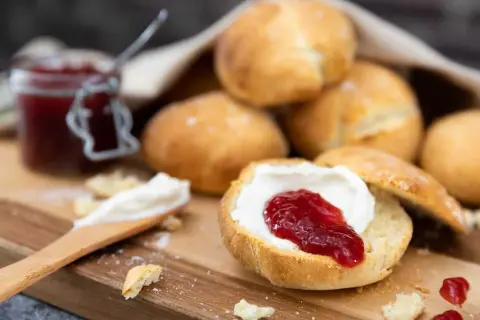 En sked med grädde och sylt på en halverad scones, redo att avnjutas. En burk sylt och fler av dessa bästa scones i bakgrunden.