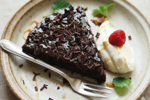 En bit kladdkaka toppad med chokladspån, serverad med vispad grädde, hallon och mynta. Perfekt för den som vill ha glutenfria kladdkakemuffins.