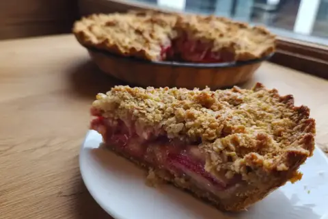 En bit av en härlig rabarberpaj med havregryn som topping. Hela pajen syns i bakgrunden.