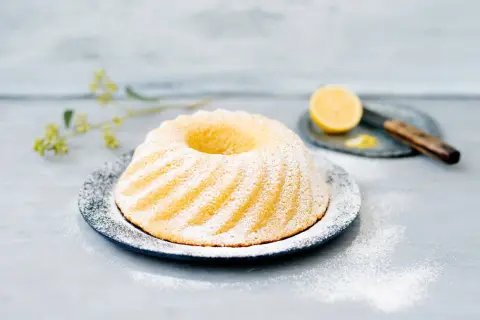 En vackert pudrad sockerkaka citron på ett fat, med en citronskiva och kniv i bakgrunden.