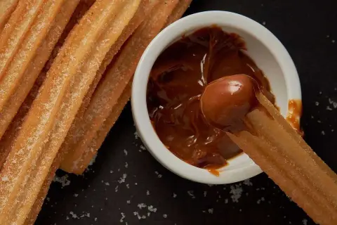 Churros coated in sugar being dipped into a bowl of dulce de leche.