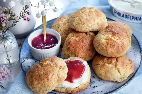 Färska scones bikarbonat på ett fat med sylt och grädde. En vacker bukett blommor i bakgrunden.