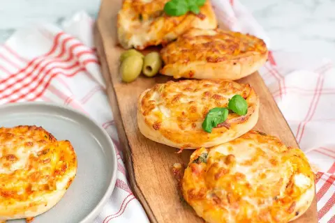 Four golden-brown pizza buns topped with melted cheese and basil, served on a wooden board with olives.