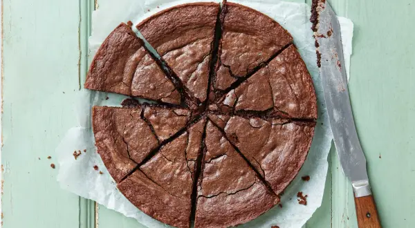 En stor, nybakad kladdkaka, uppskuren i åtta bitar, ligger på bakplåtspapper. En kniv med trähandtag ligger bredvid.