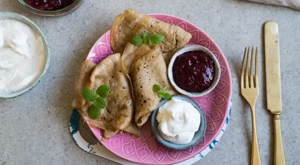 Glutenfria pannkakor med sylt och grädde på en rosa tallrik. En gaffel och kniv ligger bredvid.