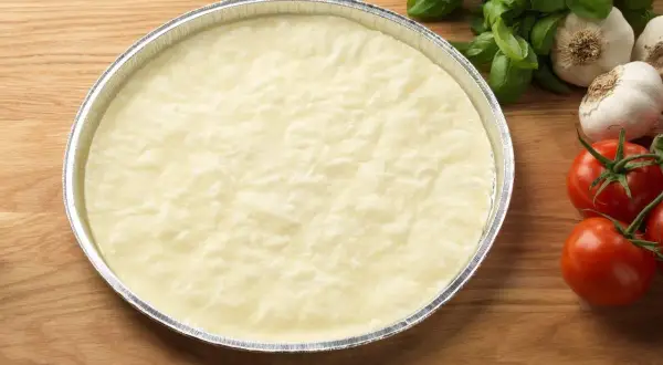 En rund, ljus deg i en folieform, redo för ett glutenfri pizzadeg recept. Tomater och vitlök i bakgrunden.