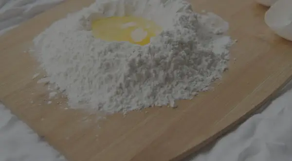 En hög mjöl med ett ägg i mitten, redo att blandas. Kanske lite för mycket bakpulver i receptet? Äggskal ligger bredvid.