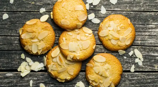 Små, gyllene mandelkakor, perfekta för ett bondkakor recept. Spridda mandelflagor på en rustik träyta.