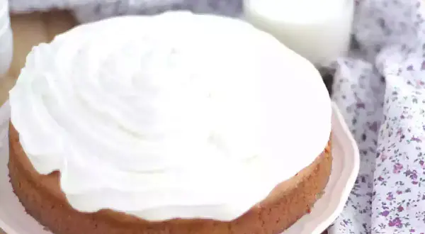 En saftig tres leches-kaka med fluffigt vitt täcke, bredvid ett glas mjölk och en blommig duk.