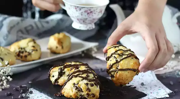En hand plockar upp en chokladscone, dekorerad med chokladglasyr. Fler chokladscones ligger på ett fat.
