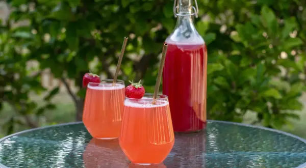 Två glas med rosa jordgubbssaft, garnerade med färska jordgubbar och sugrör, står på ett glasbord. En flaska med jordgubbssaft står bakom. Ett perfekt jordgubbssaft recept för sommaren.