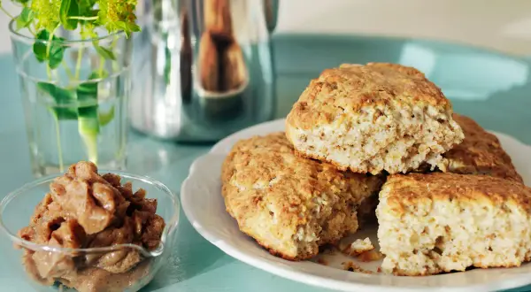 Nybakade kardemumma scones på en vit tallrik, med en skål fylld med kardemummafyllning och en vas med gröna blommor i bakgrunden.
