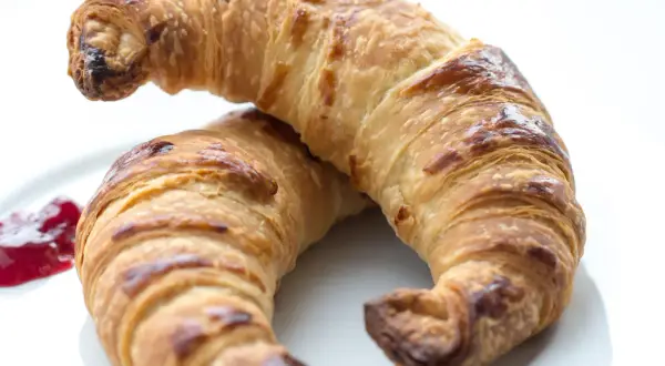 Två gyllene, krispiga croissanter på en vit tallrik med en klick sylt. Perfekt för ett croissant recept.