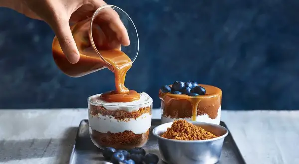En hand häller karamellsås över en läcker cheesecake i glas. Blåbär och smulor fulländar desserten.