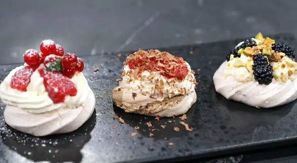 Tre små pavlovakakor med olika toppingar: jordgubbar och vinbär, choklad och hallon, samt björnbär och pistagenötter.