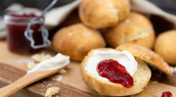 En sked med grädde och sylt på en halverad scones, redo att avnjutas. En burk sylt och fler av dessa bästa scones i bakgrunden.