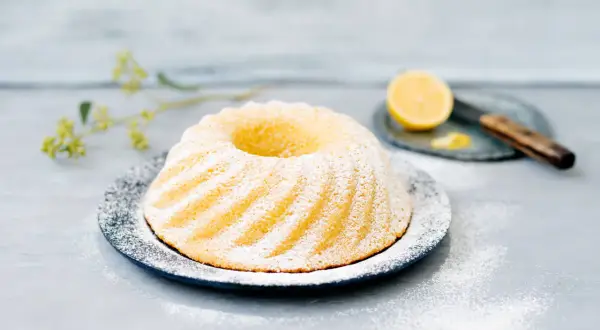En vackert pudrad sockerkaka citron på ett fat, med en citronskiva och kniv i bakgrunden.