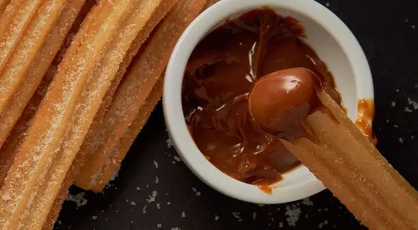 Churros coated in sugar being dipped into a bowl of dulce de leche.