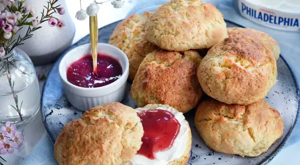 Färska scones bikarbonat på ett fat med sylt och grädde. En vacker bukett blommor i bakgrunden.
