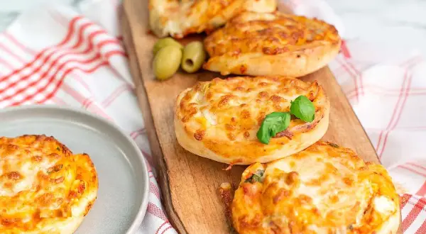Four golden-brown pizza buns topped with melted cheese and basil, served on a wooden board with olives.