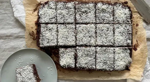 En plåt med chokladkakor, toppade med kokosflingor, redo att avnjutas som kärleksmums på påsk. En bit ligger på en tallrik.