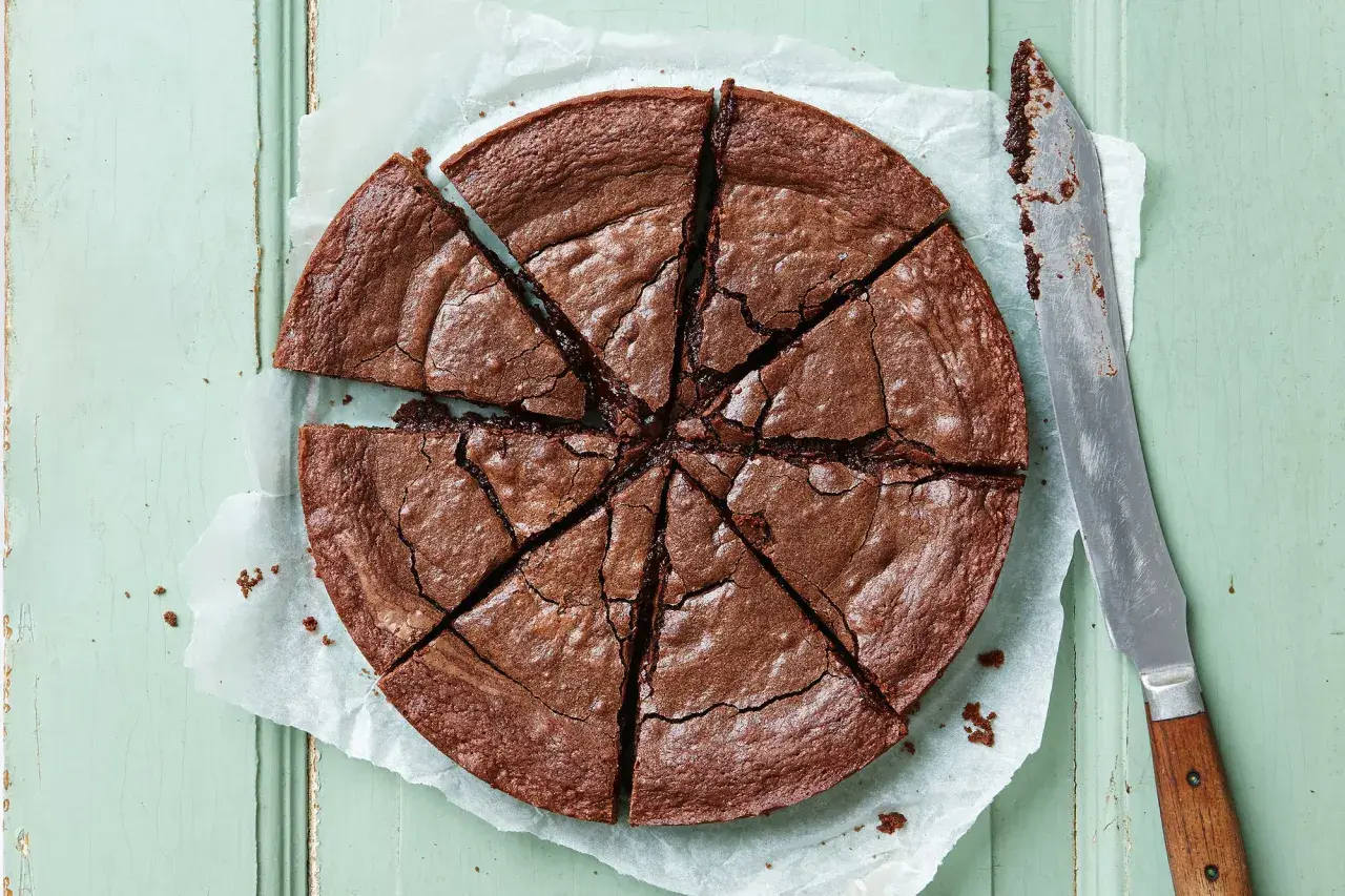En stor, nybakad kladdkaka, uppskuren i åtta bitar, ligger på bakplåtspapper. En kniv med trähandtag ligger bredvid.