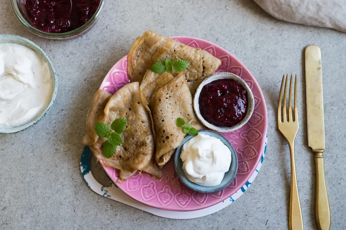 Glutenfria pannkakor med sylt och grädde på en rosa tallrik. En gaffel och kniv ligger bredvid.