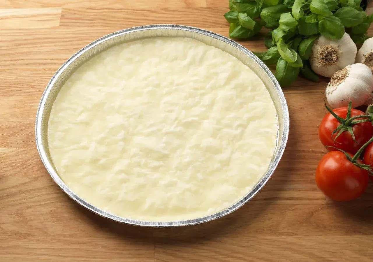 En rund, ljus deg i en folieform, redo för ett glutenfri pizzadeg recept. Tomater och vitlök i bakgrunden.