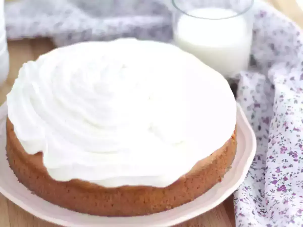 En saftig tres leches-kaka med fluffigt vitt täcke, bredvid ett glas mjölk och en blommig duk.